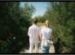 Back view of a couple walking hand in hand on a wooden bridge surrounded by greenery.