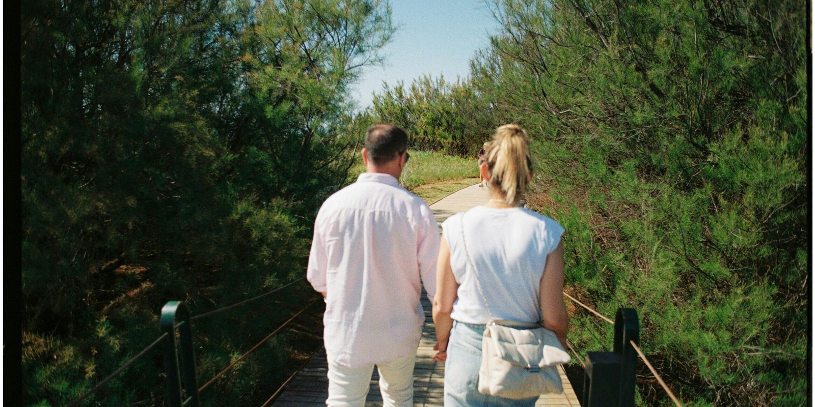 Back view of a couple walking hand in hand on a wooden bridge surrounded by greenery.