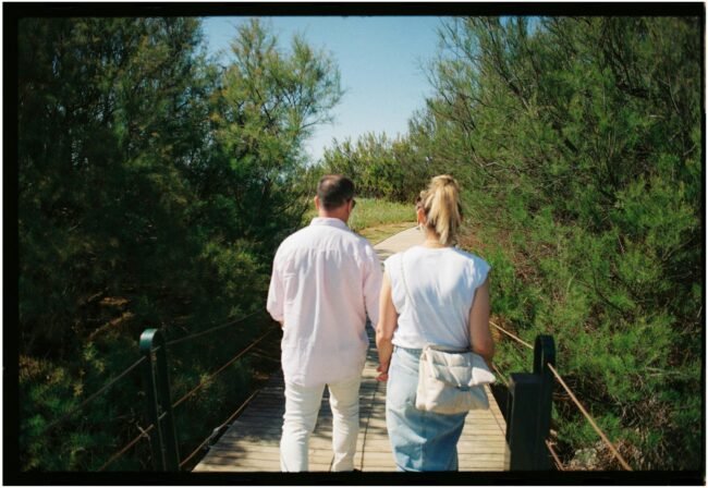 pexels-photo-23413620-23413620 Back view of a couple walking hand in hand on a wooden bridge surrounded by greenery.