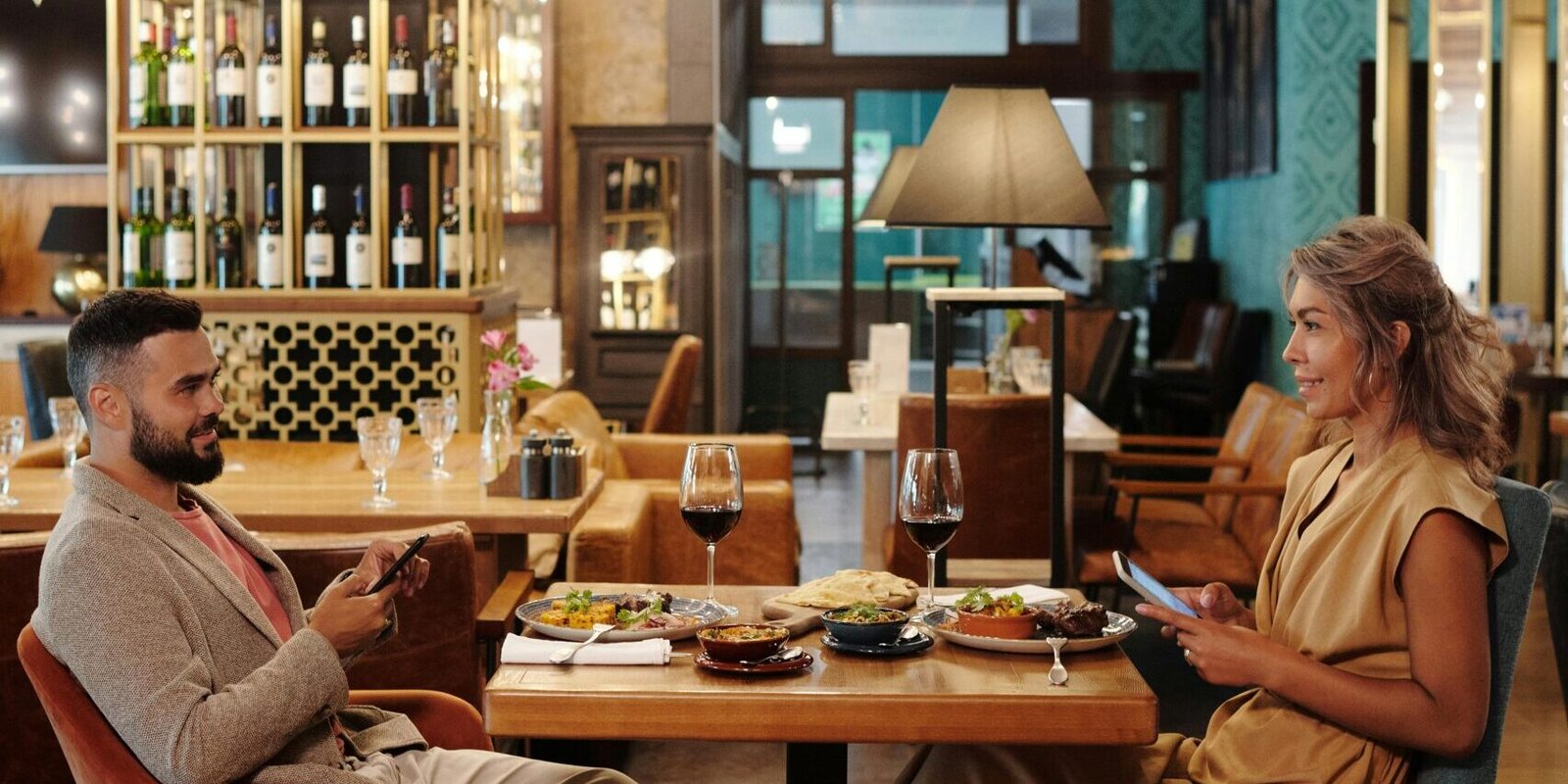 Couple enjoying a meal and conversation at a stylish restaurant, using smartphones and wine glasses present.