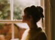A woman stands by a window holding a warm cup, looking out.