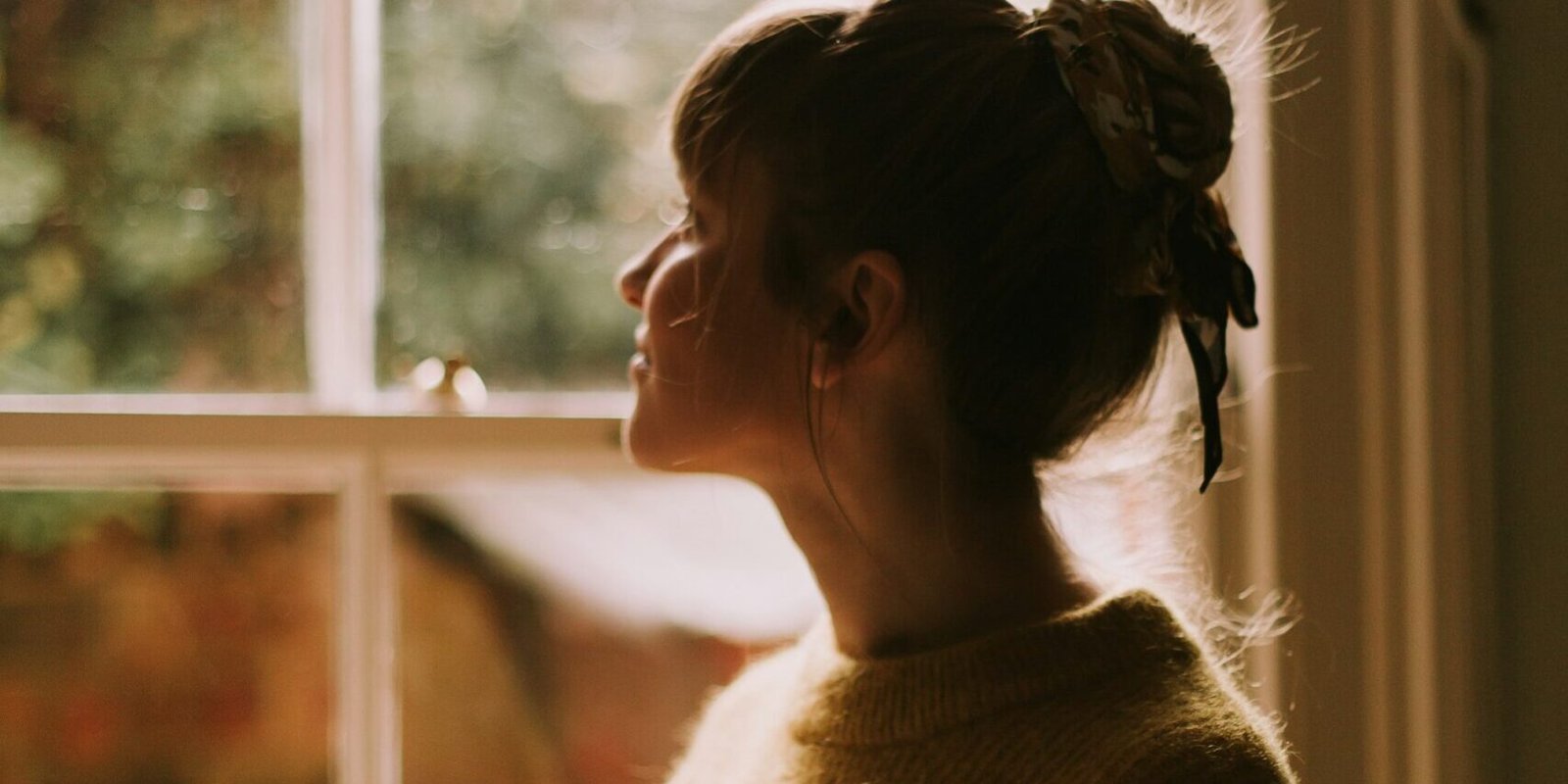 A woman stands by a window holding a warm cup, looking out.