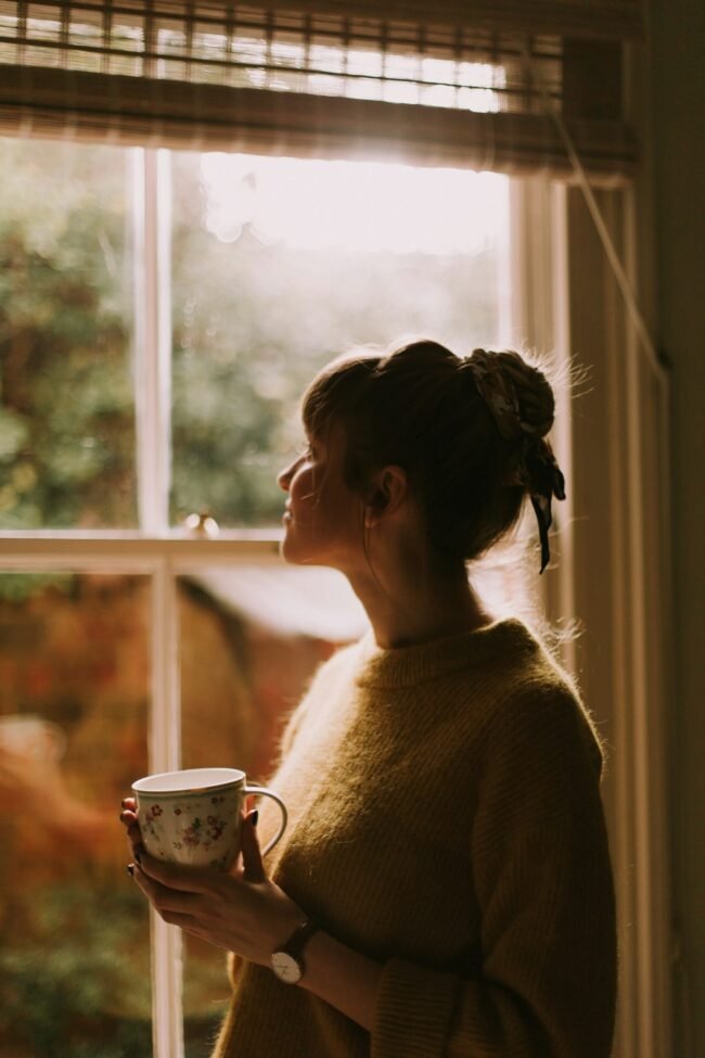 A woman stands by a window holding a warm cup, looking out.