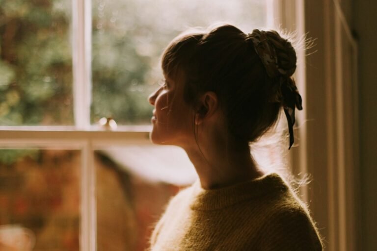 A woman stands by a window holding a warm cup, looking out.