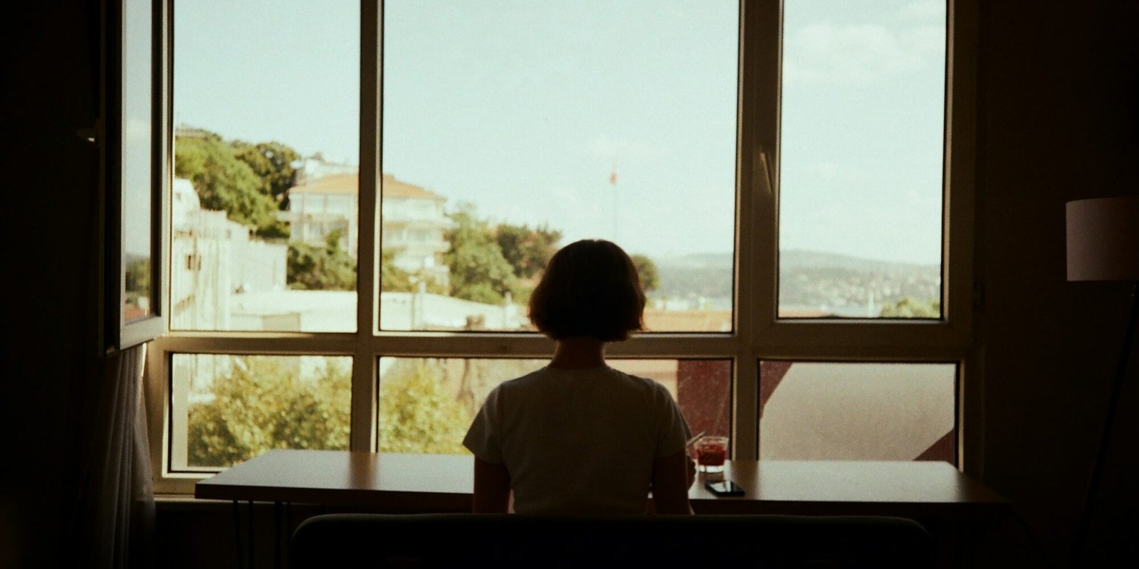A woman sitting at a table, looking out a window in soft daylight.