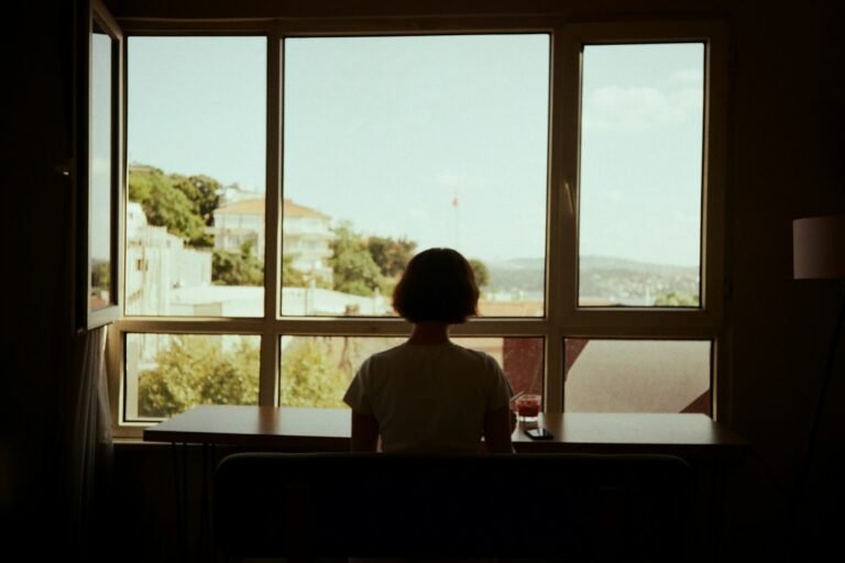 A woman sitting at a table, looking out a window in soft daylight.